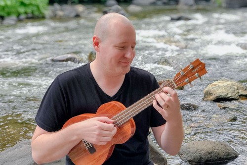 A young, happy, and vibrant Nathan Arnold playing the ukelele while sitting on a rock in an Alaska river