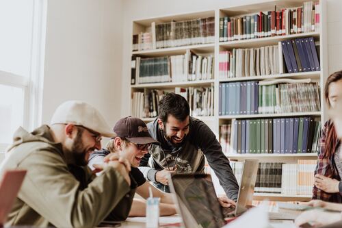 Clean Curriculum image of a small group of three males huddled around a laptop smiling and laughing with a bookcase full of books behind them.