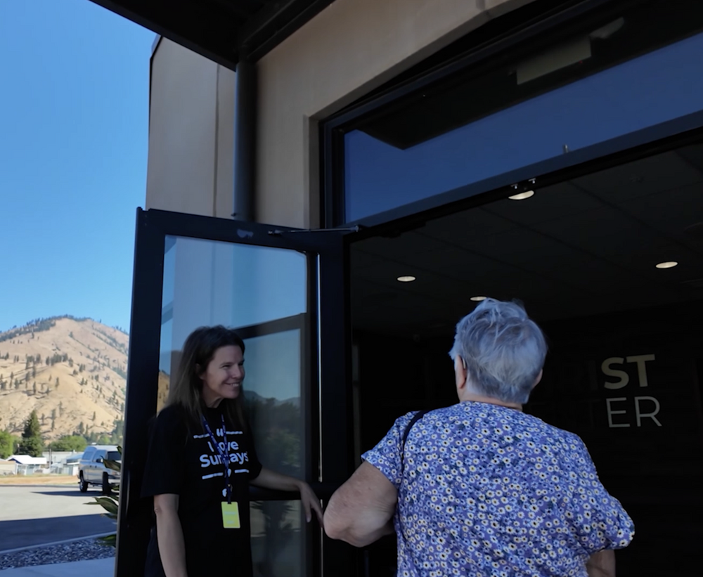 A volunteer holding the front door open and greeting a woman arriving for a service