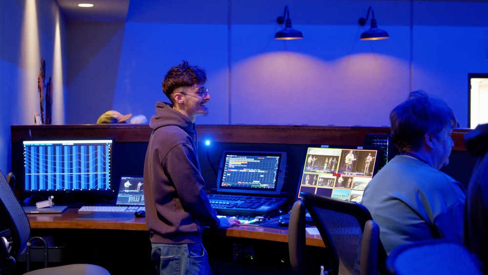 Two production volunteers at the sound and lighting desk beside the camera monitor wall