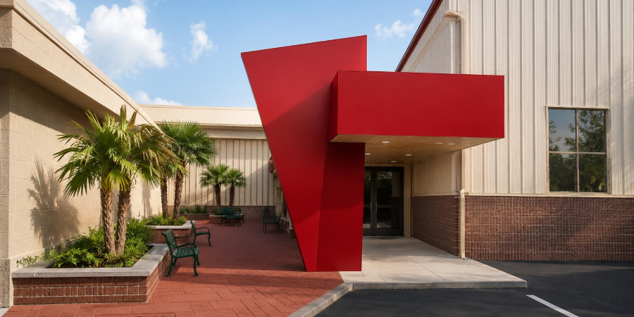 Rendering of the new multipurpose center entrance, with a red angular canopy over glass doors, palm trees and a brick paver courtyard
