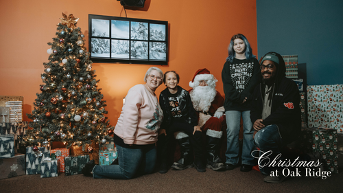 Santa Clause in a chair next to Christmas Tree with hands up waving