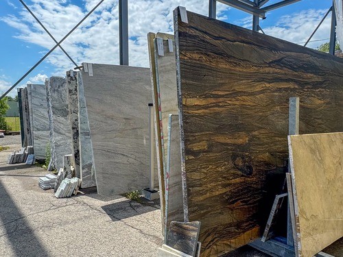 Outdoor display of various large stone slabs, including granite and marble, standing upright on supports, with a blue sky and clouds in the background.