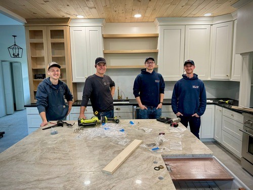 Four Red Leaf Milling Company staff members standing around a kitchen island during installation, with tools and materials on the countertop, and custom cabinetry in the background.