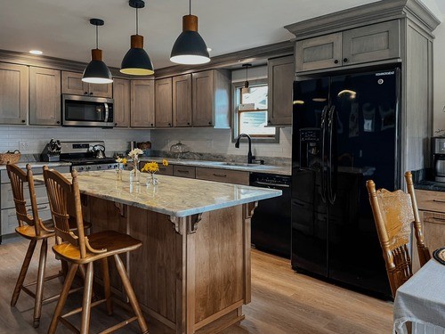 Modern kitchen with grey cabinets, black appliances, a large island with a marble countertop, wooden bar stools, and pendant lighting.