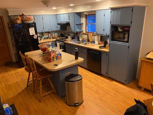 Old kitchen with blue painted cabinets, beige countertops, black appliances, a central island with bar seating, and wooden flooring.
