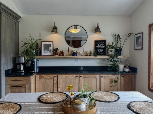 Dining area with a decorated shelf, potted plants, a round mirror, and a table set with placemats and a centerpiece on a white tablecloth.