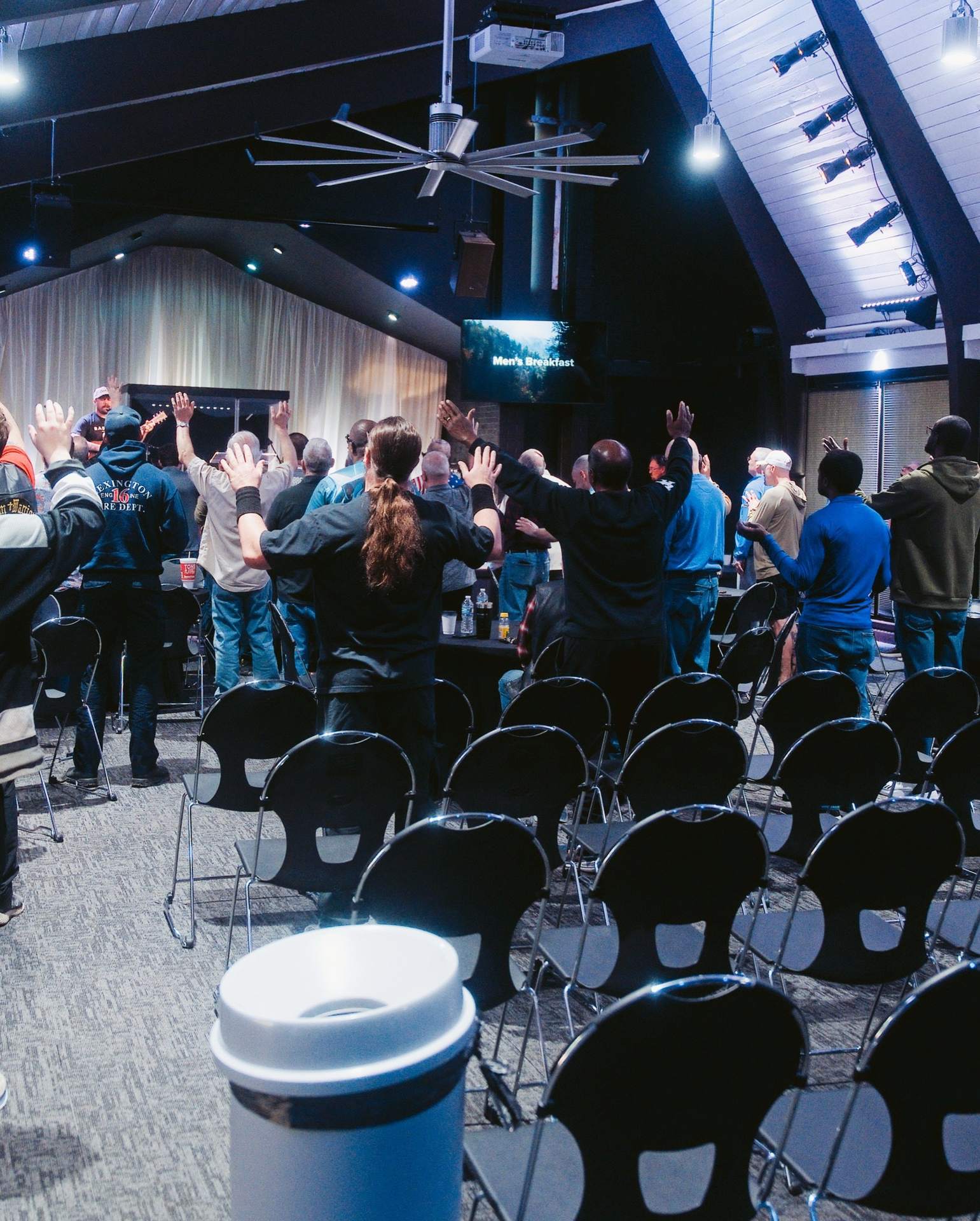 Men standing with hands raised in worship among rows of chairs