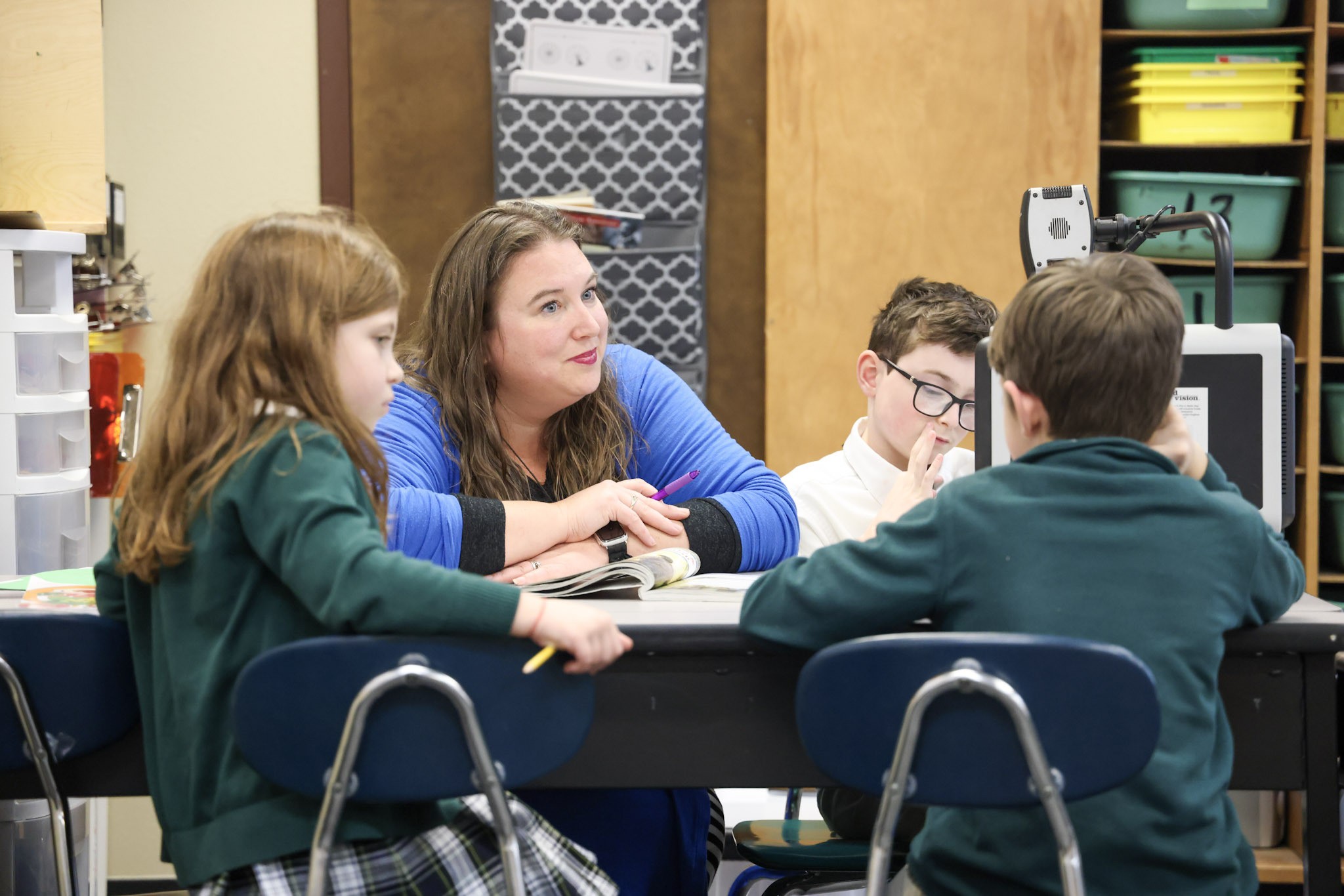 Mrs. Gaeta smiling as she gives individual attention to students around a classroom table