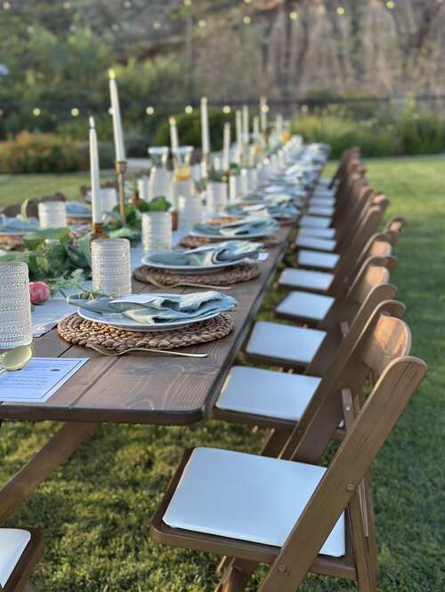 A long outdoor dining table set with woven chargers, sage napkins, glassware, and place cards