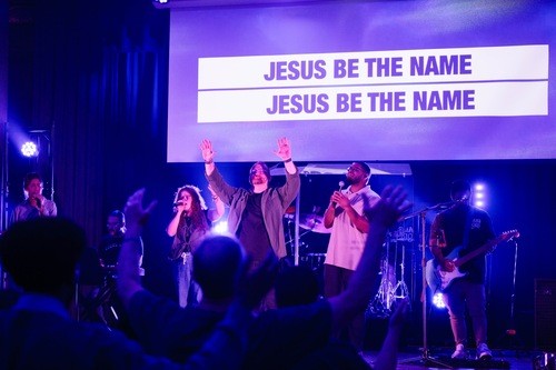 A live band performing on stage under colorful lights with the lyrics 'SINGING HOLY' displayed on a large screen behind them, while an audience watches with raised hands in the foreground.