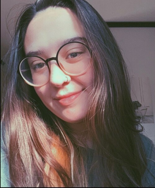 An image of Travis McFarlane, slicked back brown hair and wire-rimmed glasses, wearing a collared white shirt with light pink and blue stripes and squares with a soft smile in front of the stones in the atrium of First Christian.