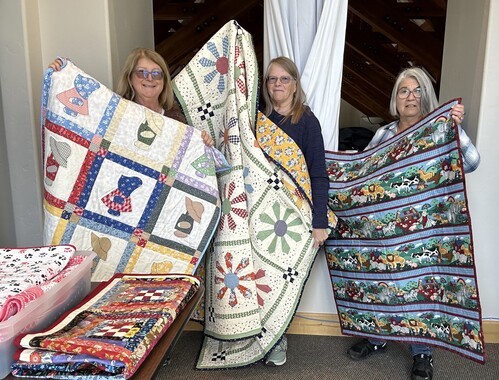 Three women showing quilts made for Habitat for Humanity homeowners
