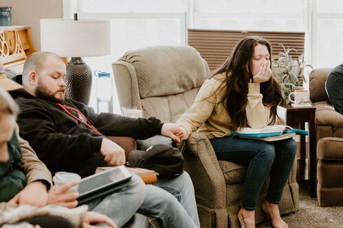 A couple sitting in chairs in a living room, eyes closed in prayer and holding hands
