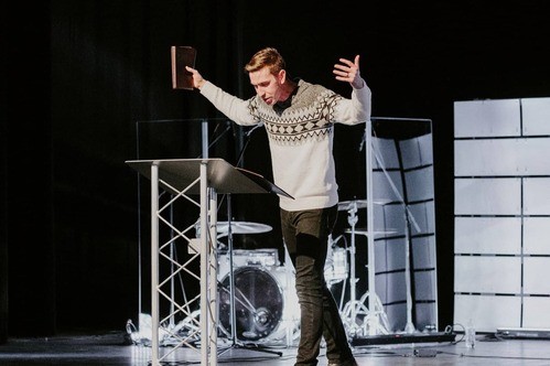 Man preaching at a podium with his arms stretched wide and a Bible in his right hand
