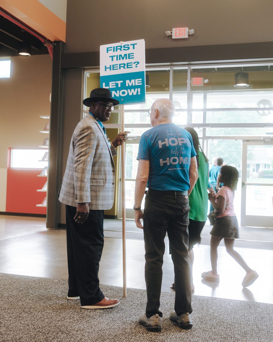 A greeter holding a First time here? Let me know! sign at the Tucker front doors as families walk in