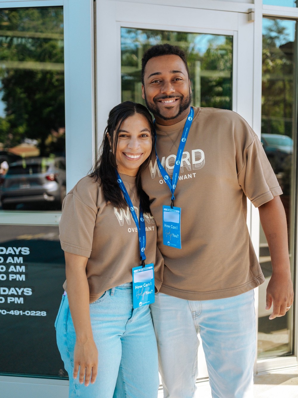 Two volunteers in How Can I Help You badges smiling together outside the Tucker campus doors