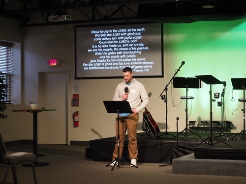 Picture of a man reading the scripture for the day, from a music stand in front of the stage, with the words of the scripture overhead projected on a screen.