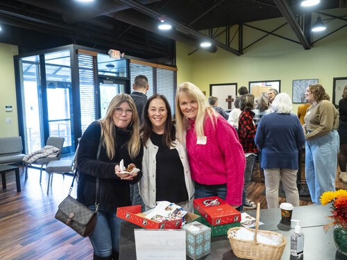 Picture of 3 women smiling as they share some pastry in the church lobby. Their are7 or 8 others also mingling in the lobby.