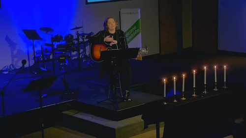 Picture of Pastor Kathryn on stage with guitar, during a dark, candle-lit service.