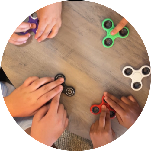 The image shows several hands of children holding and spinning different colored fidget spinners on a wooden table. The fidget spinners include colors like green, black, red, white, and purple, and the activity suggests play and engagement.