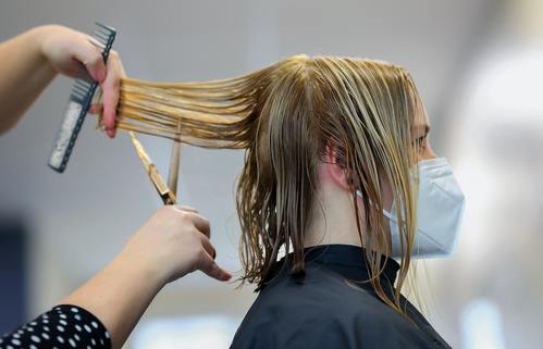 Woman is sitting in salon chair with black salon cape, and a stylist is holding up back pieces of client's hair and trimming it.